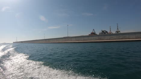 Harbor-wall-with-cranes-on-the-top-seen-from-motorboat-that-borders-the-outside,-the-blue-sea-with-the-sky-and-the-wake-of-the-boat