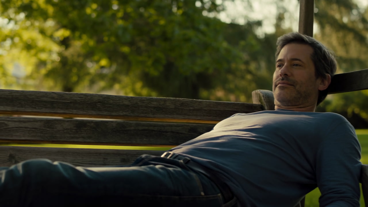 Middleaged White Man Reclining On Bench In Sunlit Park, Wearing Sweater And Jeans, Eyes Halfclosed