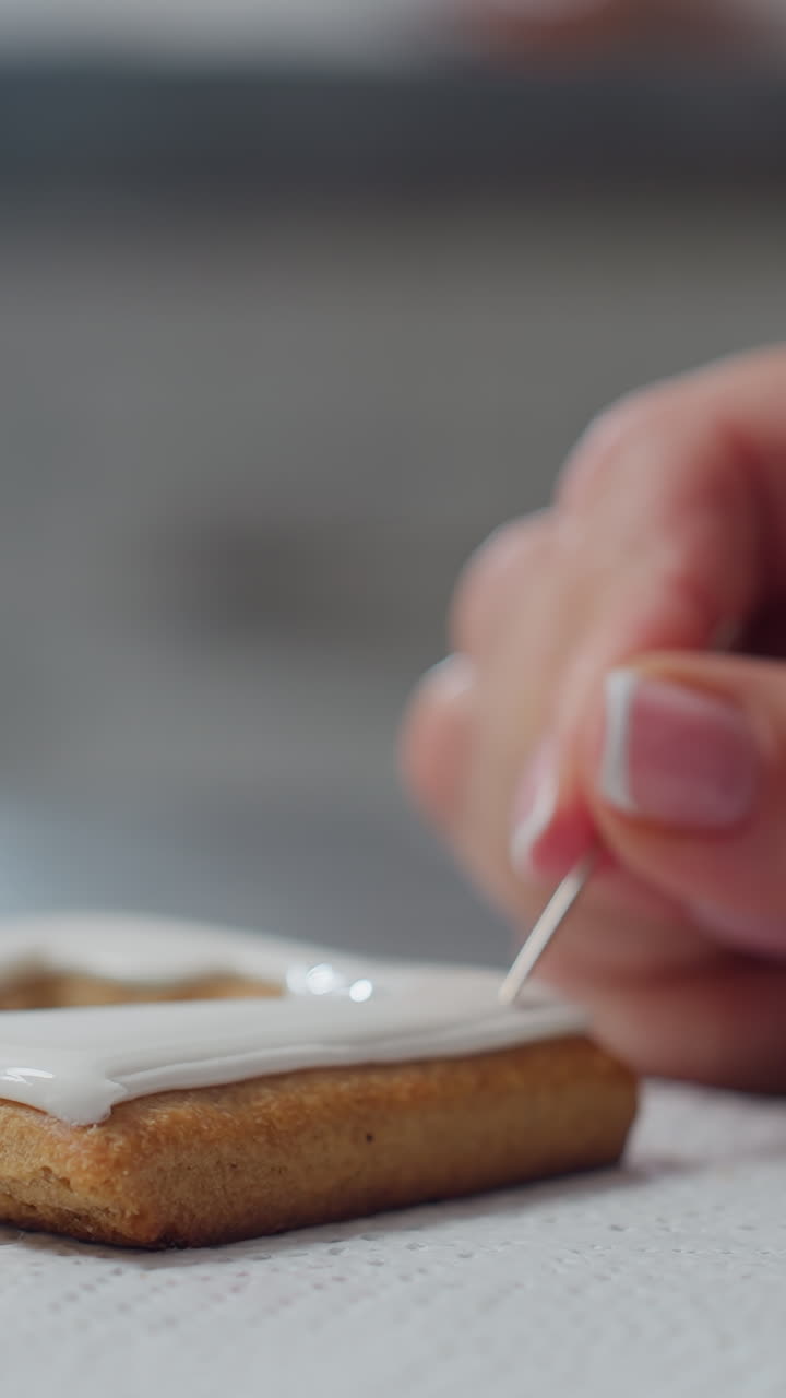Close-up of cookie with white icing being designed with toothpick held by baker, delicate decoration in progress, creating intricate designs with icing on baked goods with blurred background