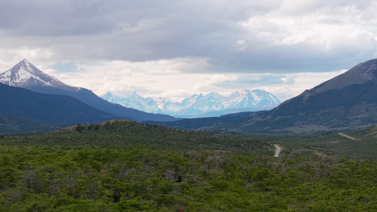 Vista distante delle cime delle Torres del Paine nella Patagonia cilena, incorniciate da montagne scure e dalla boscaglia verde con una strada tortuosa sotto nuvole suggestive