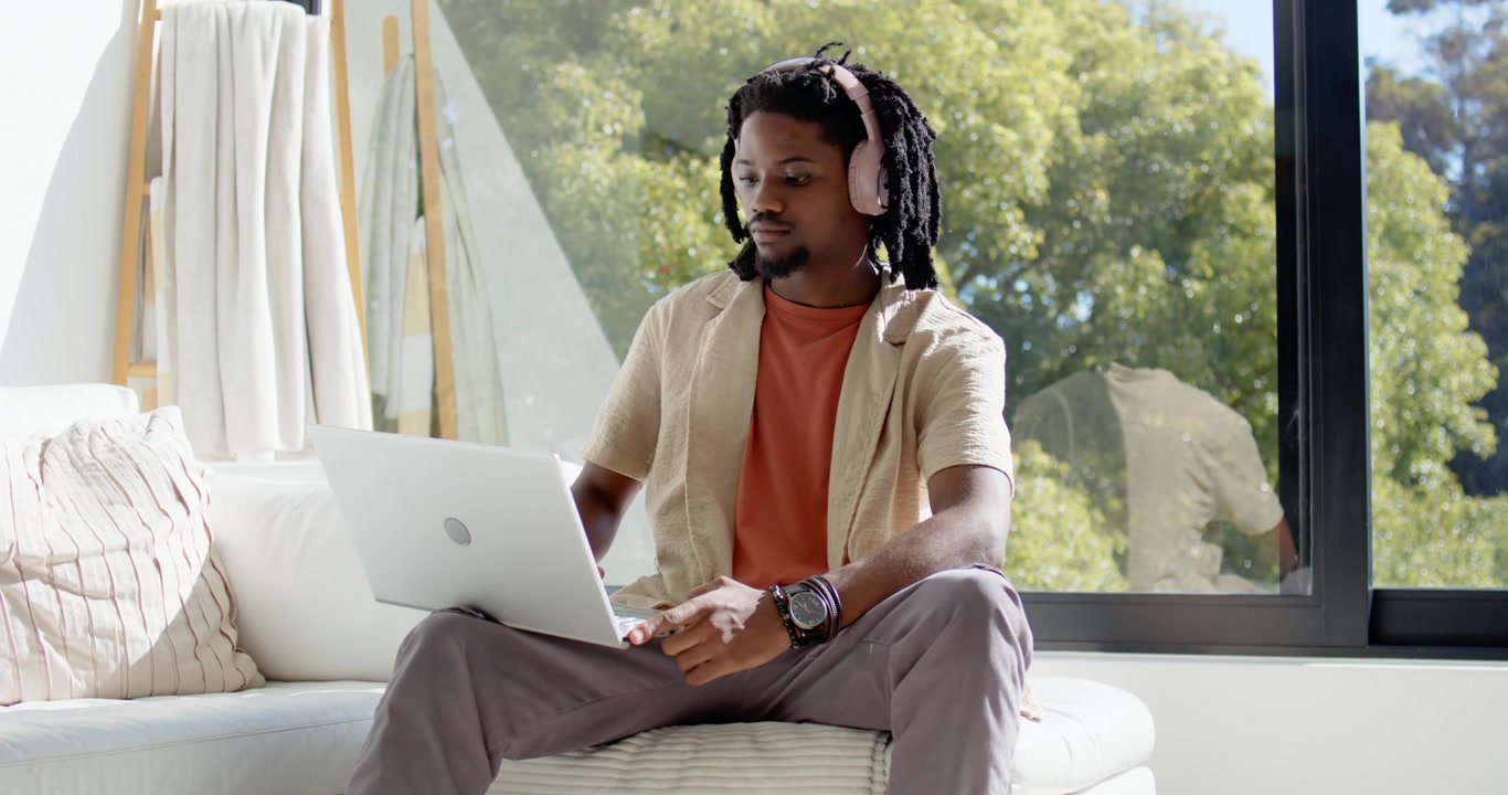 African American man wearing headphones working on silver laptop, typing, leaning on sofa by window