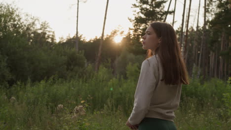 a young woman stands in a forest at sunset, looking away from the camera.