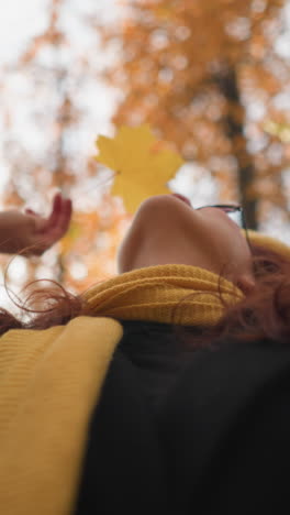 elegant lady in yellow beret and muffler holds vibrant autumn leaf over face, shielding from sunlight while gazing up at tall trees with golden foliage