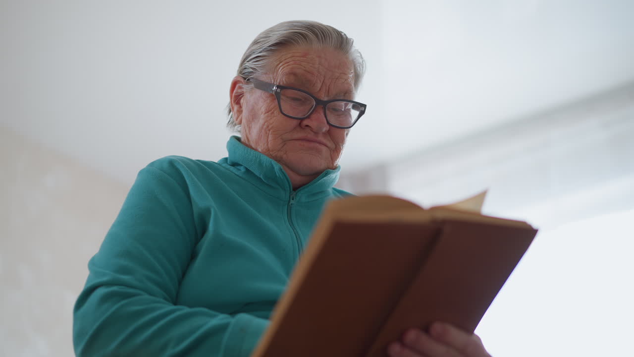 Lonely pensioner in green teal sweater reading an old brown book, engaged in a quiet moment. Indoor setting with soft natural light, creating a peaceful and reflective atmosphere
