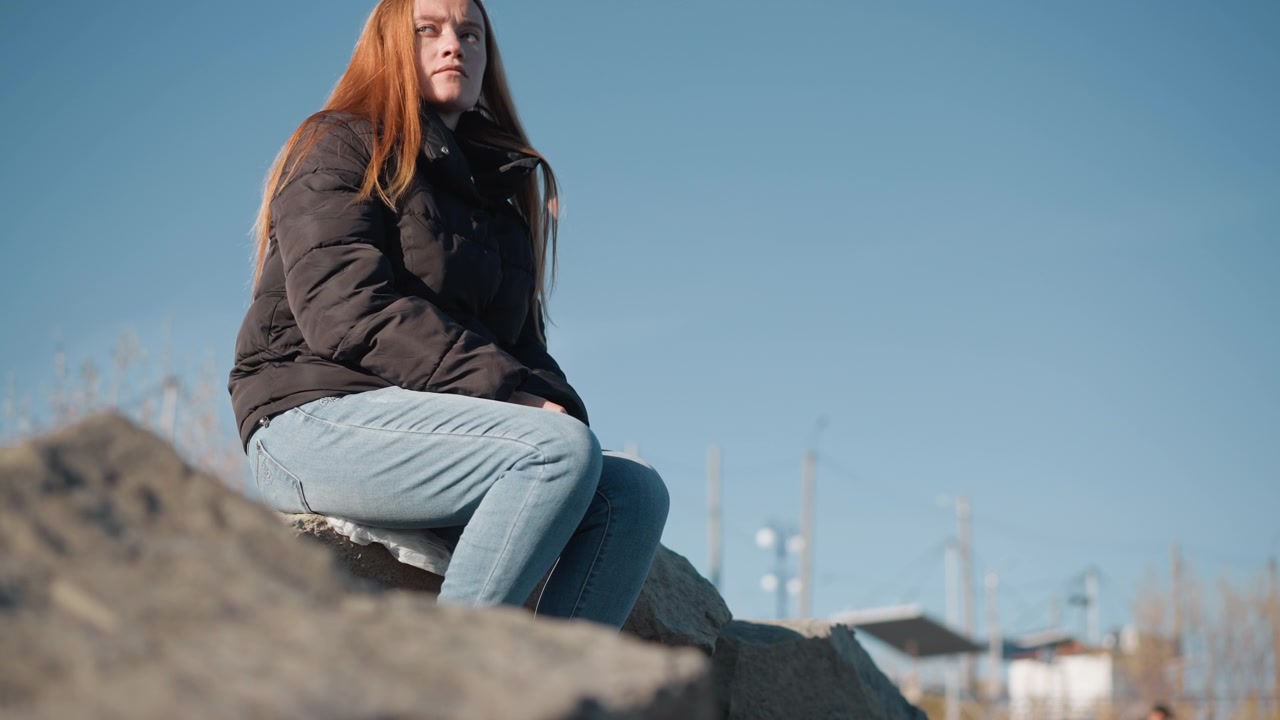 Ung kvinde sidder på en sten udendørs under en klar blå himmel, iført mørk jakke og lyse jeans. Kvinden forbliver siddende med minimal bevægelse gennem hele scenen, omgivet af naturlige elementer.