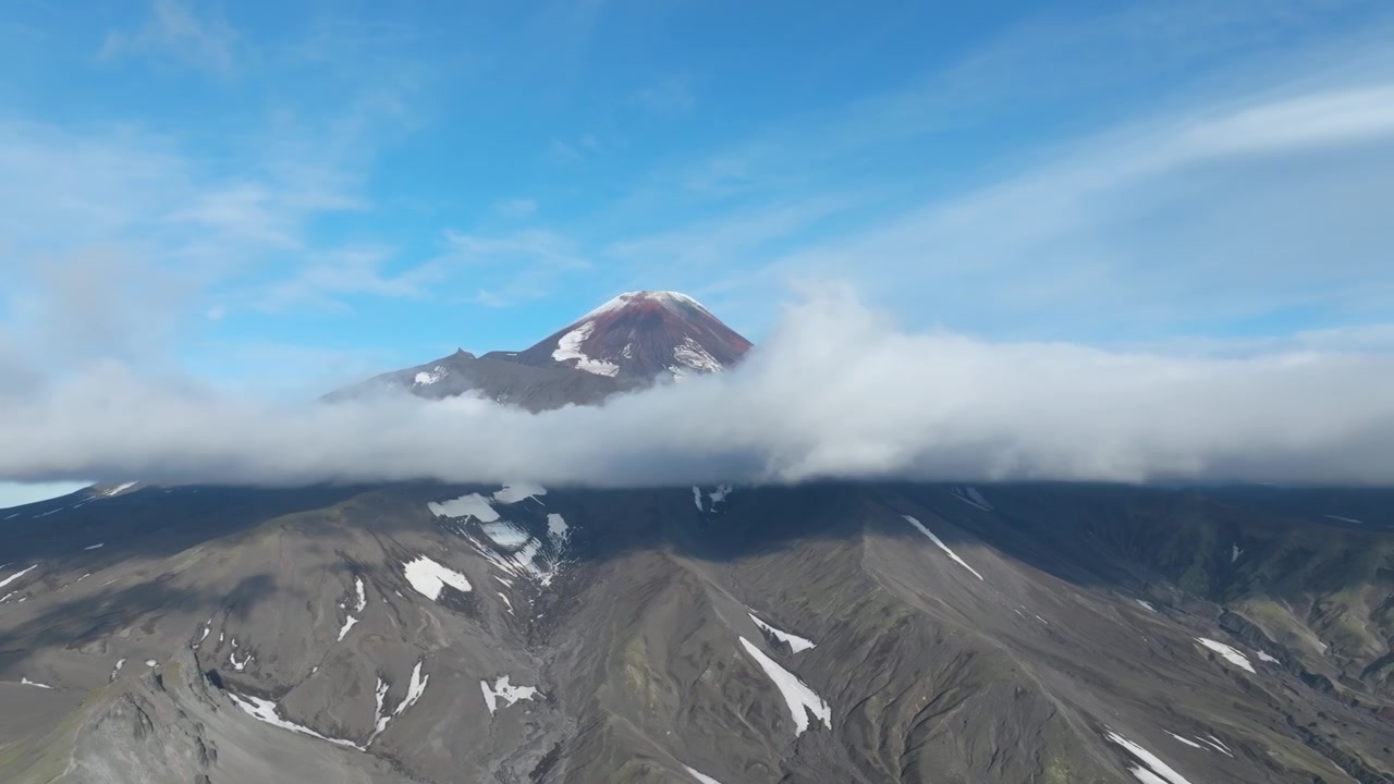 Filmato aereo del vulcano Avachinsky con uno strato di nuvole che avvolge i pendii, un paesaggio vulcanico aspro e chiazze di neve sotto un cielo blu, Territorio della Kamchatka, Russia.