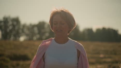 close up of woman smiling as she walks outdoors with pink shirt blowing in wind on sunny day, enjoying fresh air in vast farmland with distant trees in background