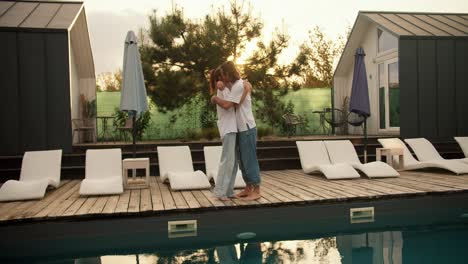 un tipo y su novia de pie en camisetas blancas cerca de las camas de sol junto a la piscina y abrazar en el fondo de la puesta de sol. descanso en la casa de campo