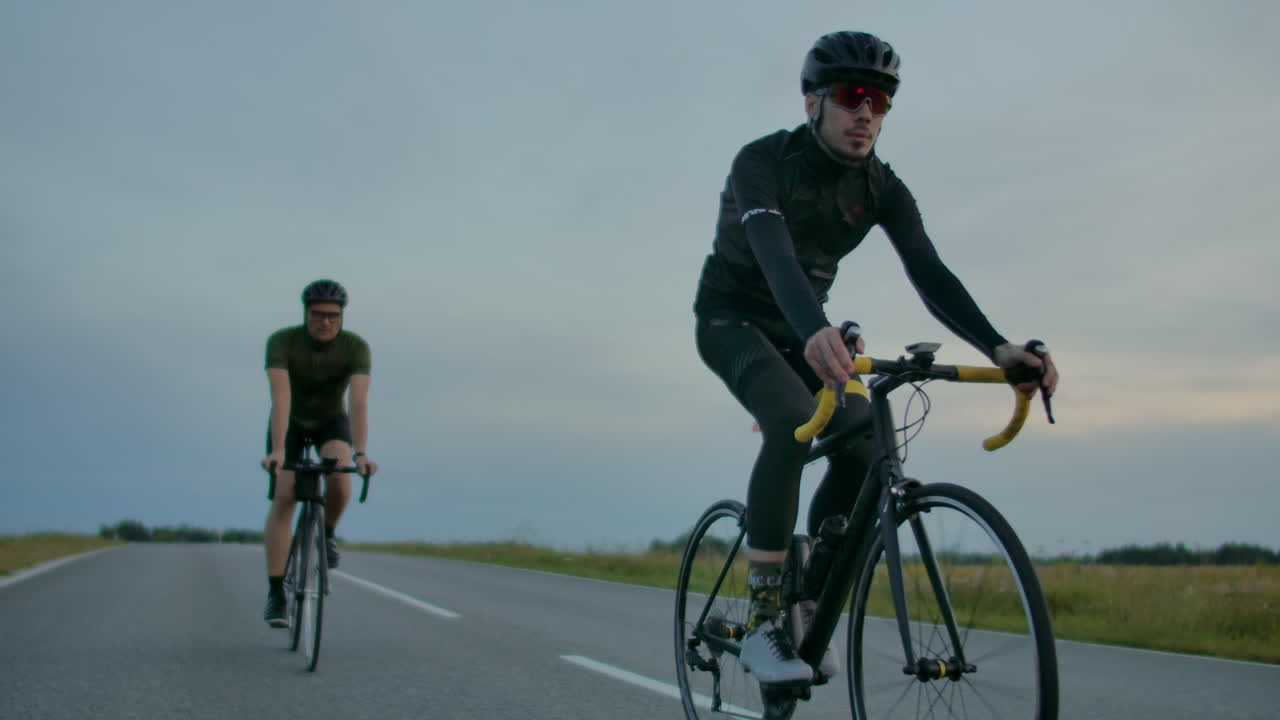 Handsome bearded professional male cyclist riding his racing bicycle in the morning together with his girlfriend both wearing protective helmets and eyeglasses sun shining through between them