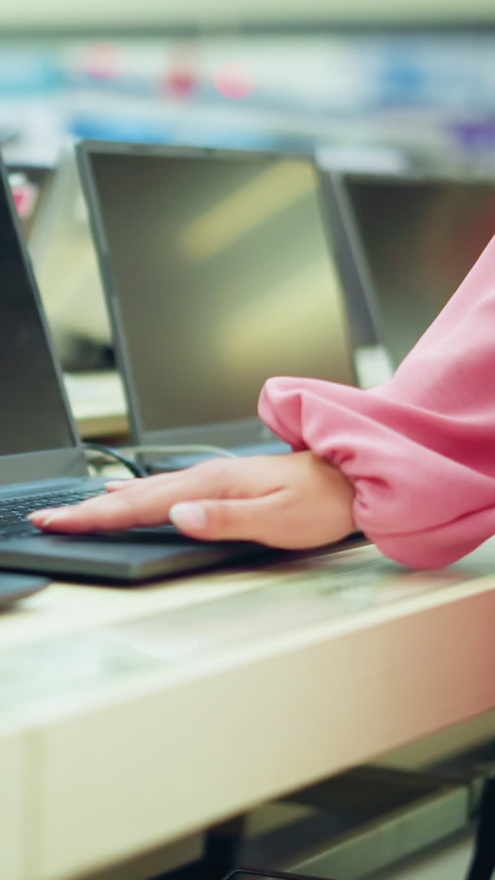 vista del cuerpo de la dama en vestido rosado con bolso negro interactuando con una computadora portátil en exhibición en una tienda de tecnología brillante, la pantalla de la computadora portable está en blanco mientras la opera, con otras computadoras portátiles alineadas
