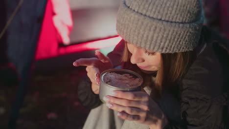 close up of young lady squatting outdoors near camp tent taking bite of can food smiling casual clothing beanie forest setting evening light natural environment camping happy moment