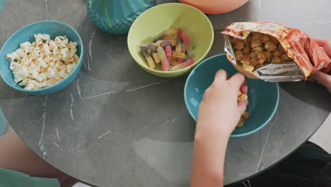 overhead view of person putting flavored popcorn back into pack while friend picks popcorn that fell on table, casual kitchen setting, snack time, friendship moment, relaxed atmosphere