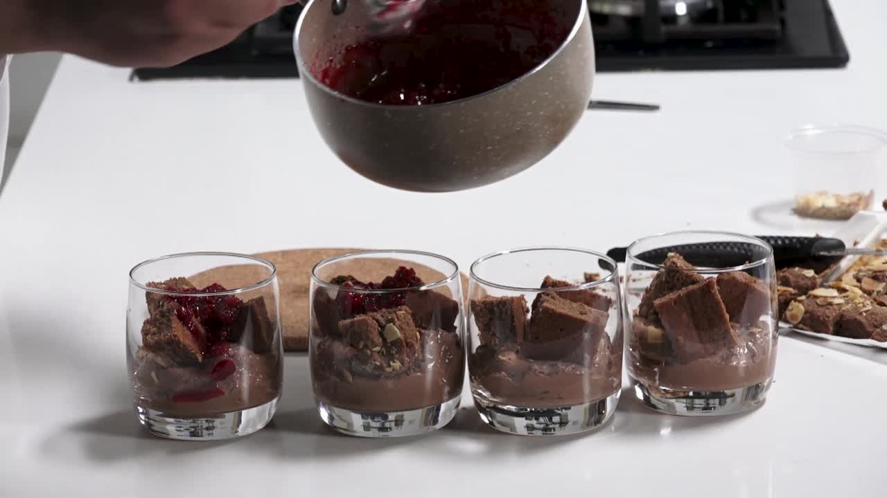 A chef skillfully layers chocolate mousse with crumbled cake and adds a vibrant berry sauce on top. This mouthwatering dessert is made in a modern kitchen