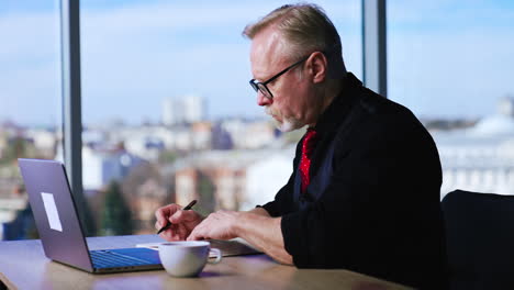 bearded man in glasses types at his laptop. adult entrepreneur taking notes in his paper book. blurred backdrop.