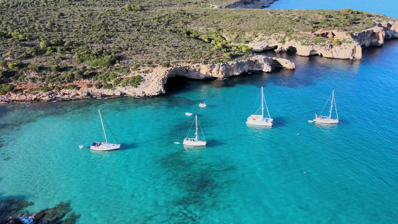Experience the stunning beauty of Cala Varques, located near Porto Cristo in Majorca. Sailboats float serenely in the vibrant turquoise waters, surrounded by lush greenery and rocky cliffs.