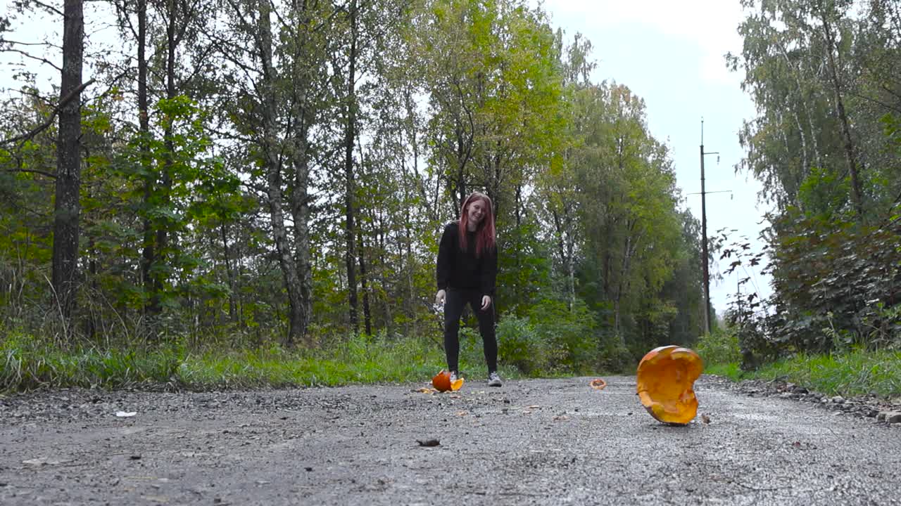 Gorgeous woman wearing black clothing throwing and destroying an orange Halloween pumpkin to the ground. Pumpkin explodes to pieces and starts to roll in slow motion. Footage filmed outdoors in autumn