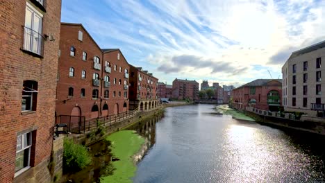 camera glides alongside a canal bordered by historic brick buildings under bright daylight, capturing reflections, greenery, and urban architecture in leeds, england