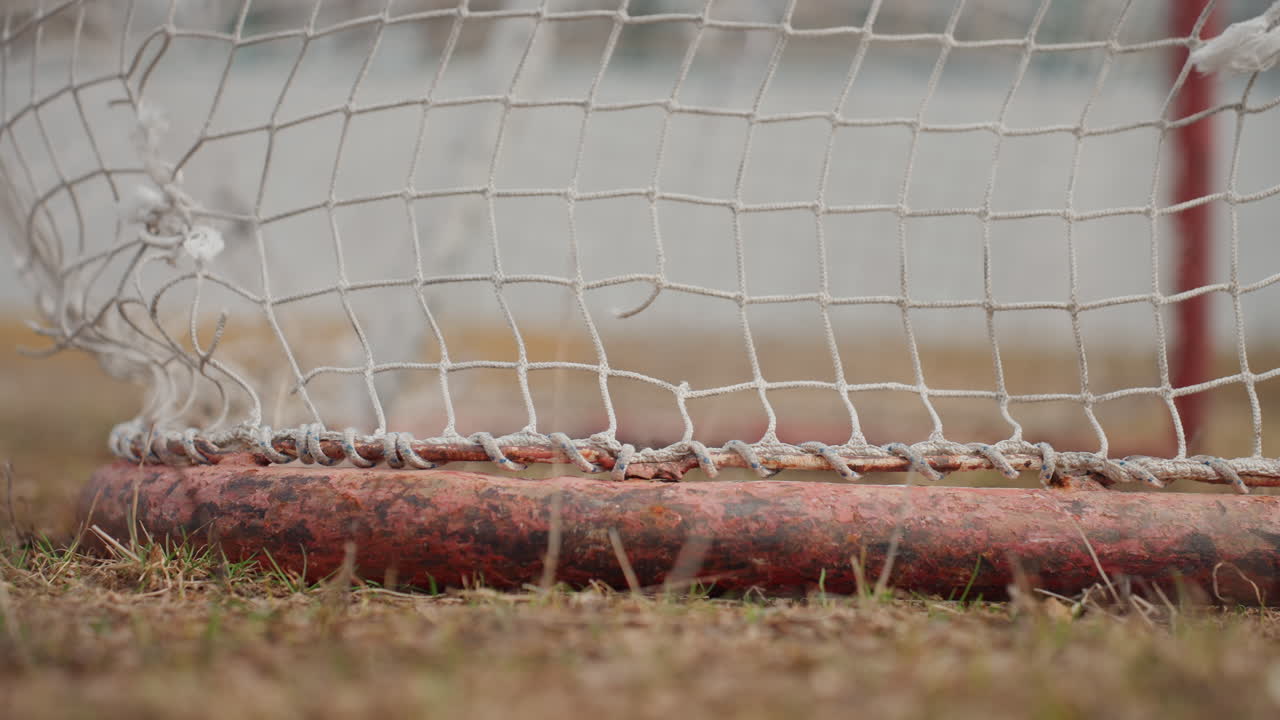 primer plano de la base de un poste de portería con red, textura áspera y pintura desgastada; estudio a cámara lenta de malla de nailon y metal oxidado, hierba seca bajo los pies, tonos otoñales apagados, textura deportiva íntima para uso editorial