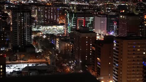 1-2 rooftop winter city semi birds eye view of post modern residential retail commercial buildings some framed in led green red lights in the late night evening with snow covered parked cars vehicles