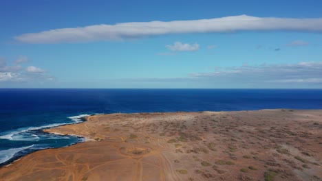 a rugged desert peninsula stretches into vivid blue ocean waves, with swirling sand tracks and a wide open sky filled with drifting white clouds.