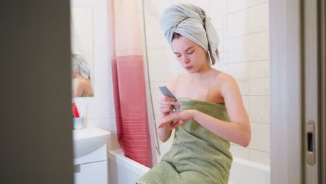 girl wrapped in light green towel, sitting on toilet, applying cream from tube on her hand, other cosmetic products visible in the bathroom background as part of her skincare routine