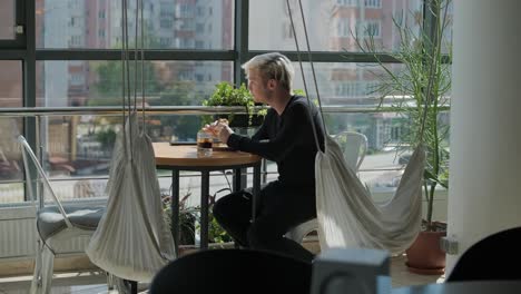 young man with blond hair enjoying a sandwich and iced coffee at a cafe table by a window with city views, natural light, and potted plants. relaxed and casual dining scene