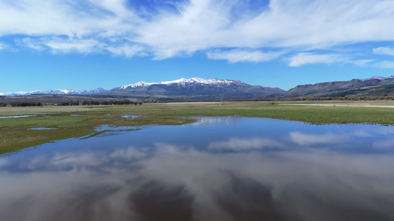 Immerse yourself in the stunning cinematic landscape of Trevelin, Argentina, featuring clouds reflecting on a pristine lake with snow-capped mountains and clear blue skies.