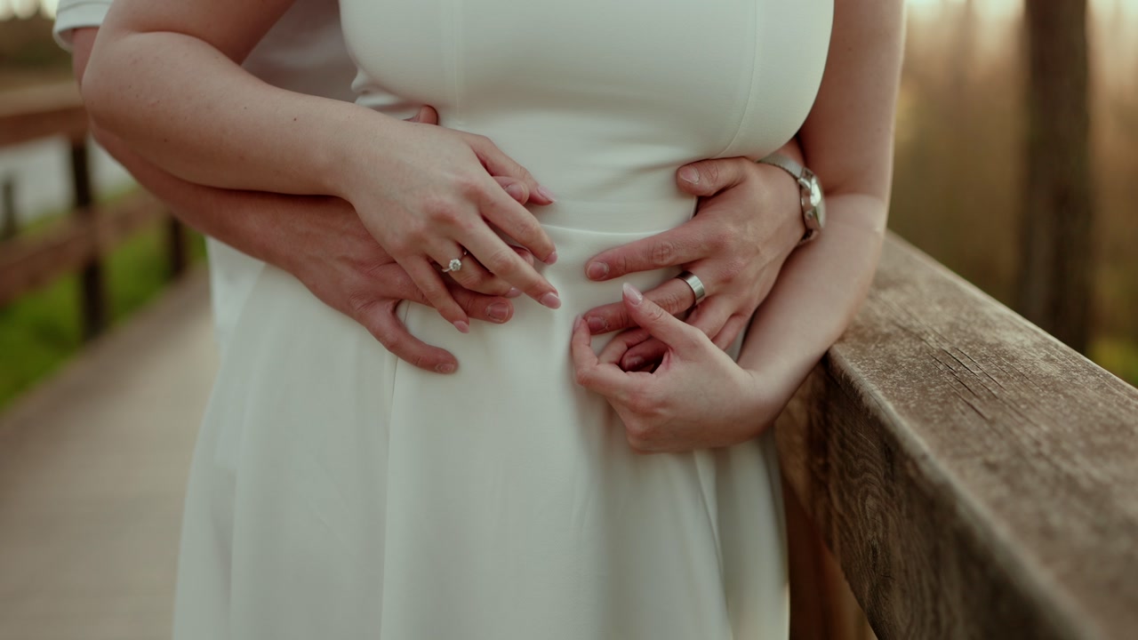 Close view of bride in white dress with partner holding her from behind while standing on wooden bridge