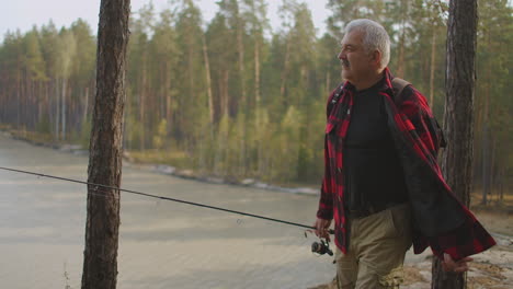 el pescador está vestido con una camisa a cuadros roja y lleva una caña de pesca giratoria caminando por la costa alta del lago