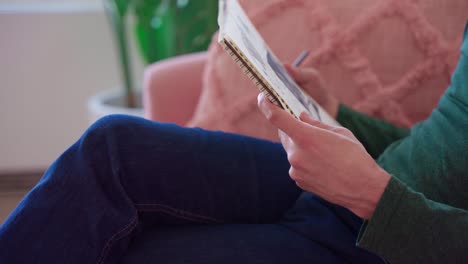 artista con camisa verde y jeans azules con cabello castaño largo dibujando la imagen de la cara de una mujer en un cuaderno de bocetos mientras se sienta en un sofá rosa