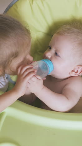 blonde girl gives to drink water from bottle to little boy sitting in soft green highchair in light room at home