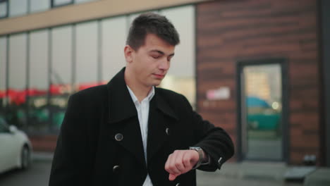 man in black coat checking time with confident look, hand in pocket, standing in front of modern glass building, successful businessman exuding professionalism in urban environment