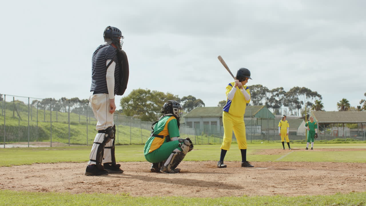 Multiracial female baseball players and male umpire playing baseball, throwing the ball on a pitch