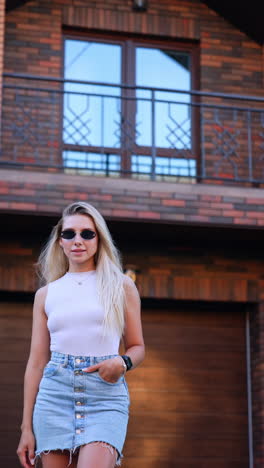 kyiv, ukraine, 25 may 2025: young woman posing outside modern house. a young woman stands confidently in front of a stylish brick house, showcasing her fashionable outfit and sunglasses