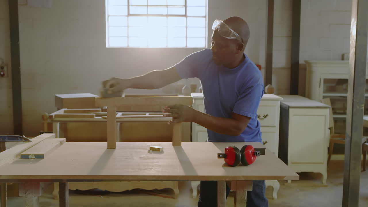 After placing bench frame, mature African American carpenter sanding with block at shop, copy space