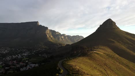 Toma-Aérea-Cinematográfica-Ascendente-Del-Pico-De-La-Cabeza-De-León-De-Ciudad-Del-Cabo-Con-La-Montaña-De-La-Mesa-Y-La-Colina-De-La-Señal-Durante-La-Puesta-De-Sol-De-La-Hora-Dorada