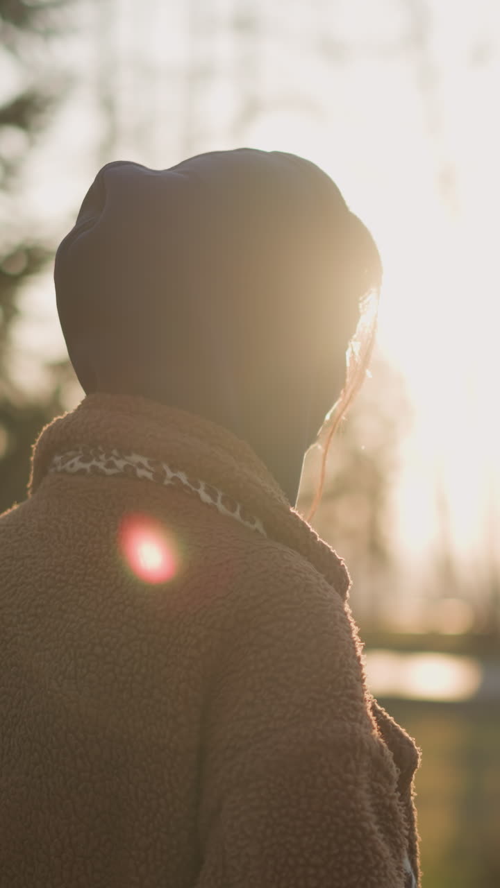 девушка идет по парку на закате, в коричневом пальто и капюшоне с капюшоном, скрывая волосы. задний вид захватывает мрачное, интроспективное настроение, когда она движется по парку