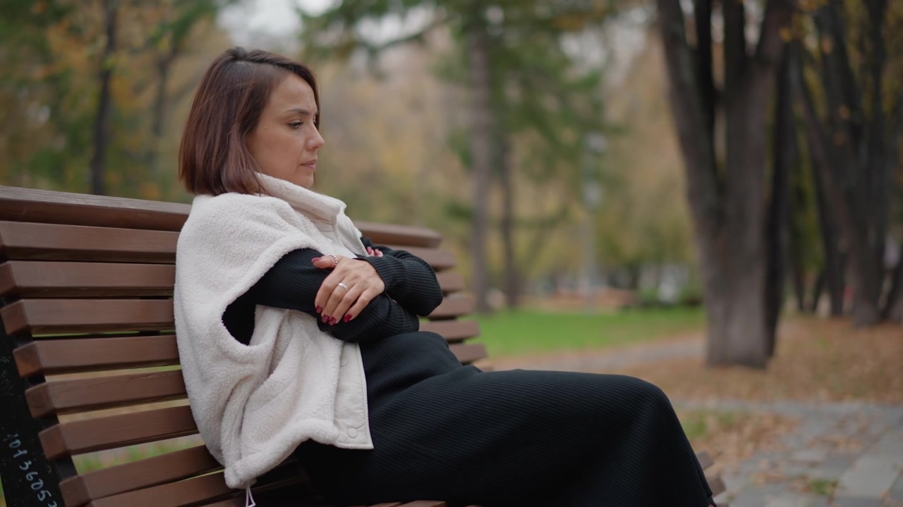 Middle aged woman sits on wooden park bench wrapped in textured knit shawl, holding it closed while turning head left, with soft autumn foliage and calm reflective mood outdoors, with woman, bench
