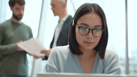 an young woman working with a laptop while two men talk in the background behind her