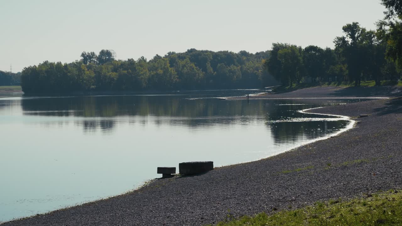 A quiet lakeside scene in soft morning light shows calm water reflecting a line of trees and a curved pebble beach, creating a serene and minimal composition