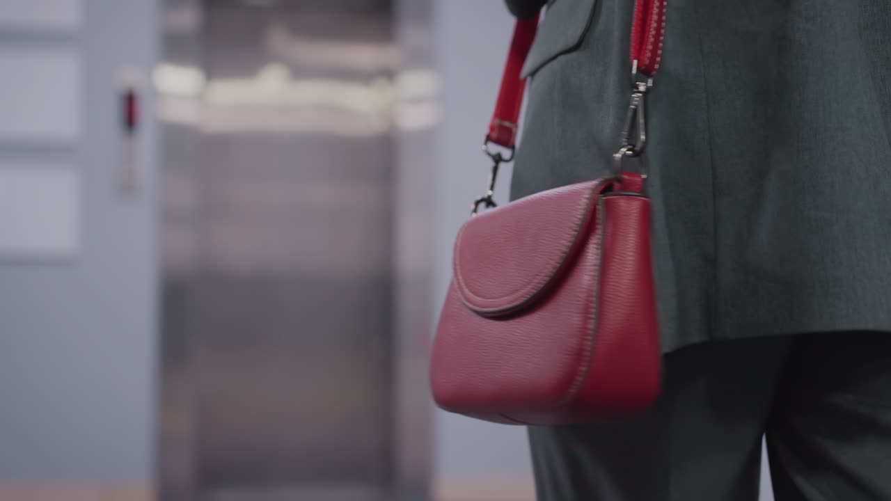 Close-up shot of red leather handbag carried by confident businesswoman in office attire. Stylish accessory complementing professional look in modern office environment, showing details of bag