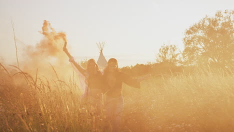 two female friends camping at music festival running through field with smoke flares