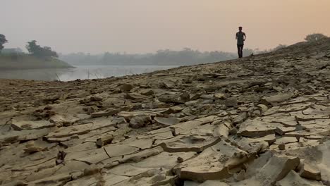 Establisher-wide-shot-of-young-man-jogging-on-dry-cracked-soil-terrain-beside-river-in-winter-monring,-Sylhet