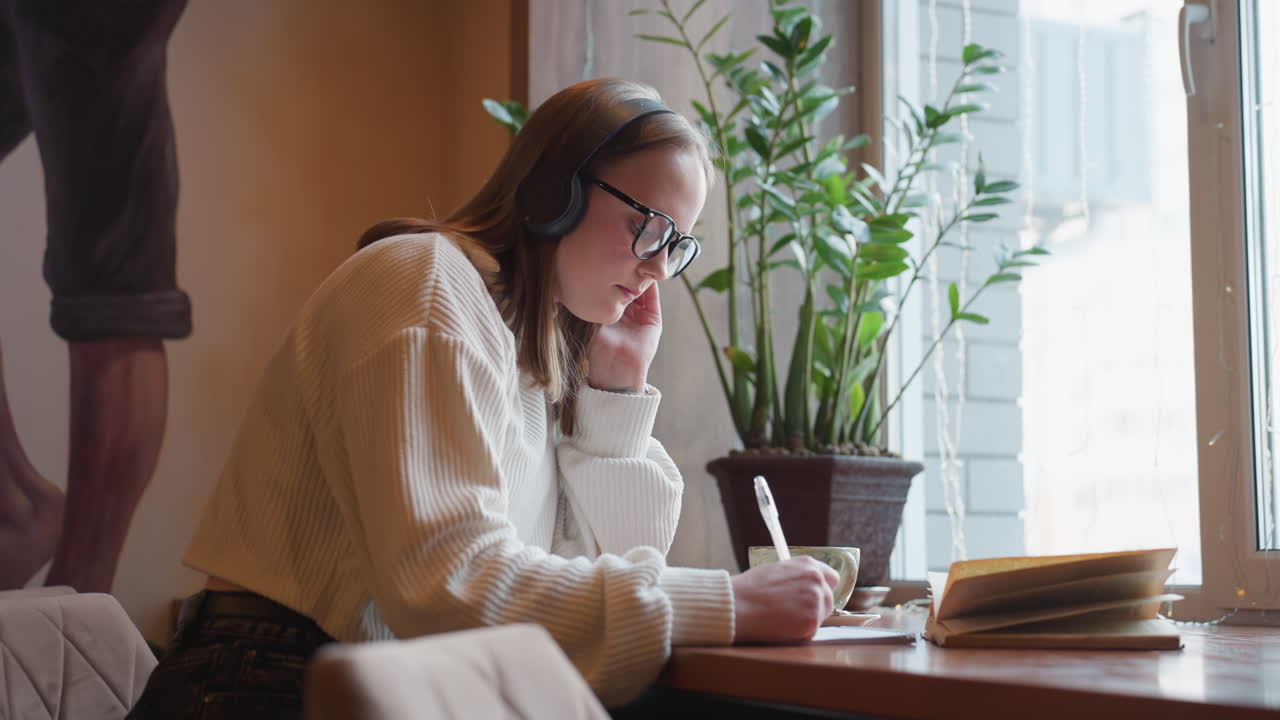young woman wearing glasses and headset writes in notebook while thoughtfully referencing open book, seated at wooden desk near window with coffee cup, ambient lighting, and indoor plant nearby