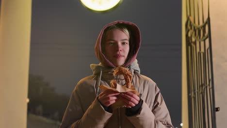 mujer joven con chaqueta de invierno con capucha de pie al aire libre por la noche, con pasteles crujientes en las manos. reloj iluminado en el fondo agrega un brillo cálido al entorno urbano fresco