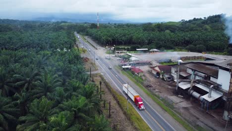 Aerial-drone-shot-from-above-a-palm-plantation,-showing-the-smoking-chimney-of-the-processing-factory-and-the-nearby-road-full-of-highly-polluting-vehicles