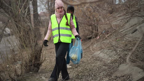 volunteers in bright safety vests walking along sloped riverbank during environmental cleanup, carrying blue plastic bags with collected litter to protect nature, reduce pollution