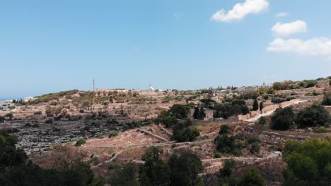 Beautiful-aerial-sideways-view-of-sunny-hills-with-green-shrubs-in-foreground-from-Top-of-the-World-in-Gharghur,-Malta