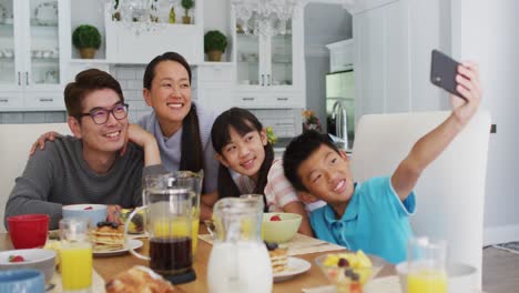 幸せなアジアの両親がキッチンで息子と娘と朝食を食べ ⁇ 息子がグループセルフィーを撮っています ⁇