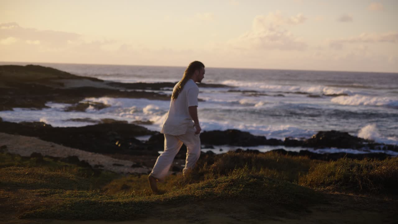 A man takes a step on a grassy bluff by the coast, gazing at ocean waves crashing against dark rocks as the golden sunset glows along the horizon